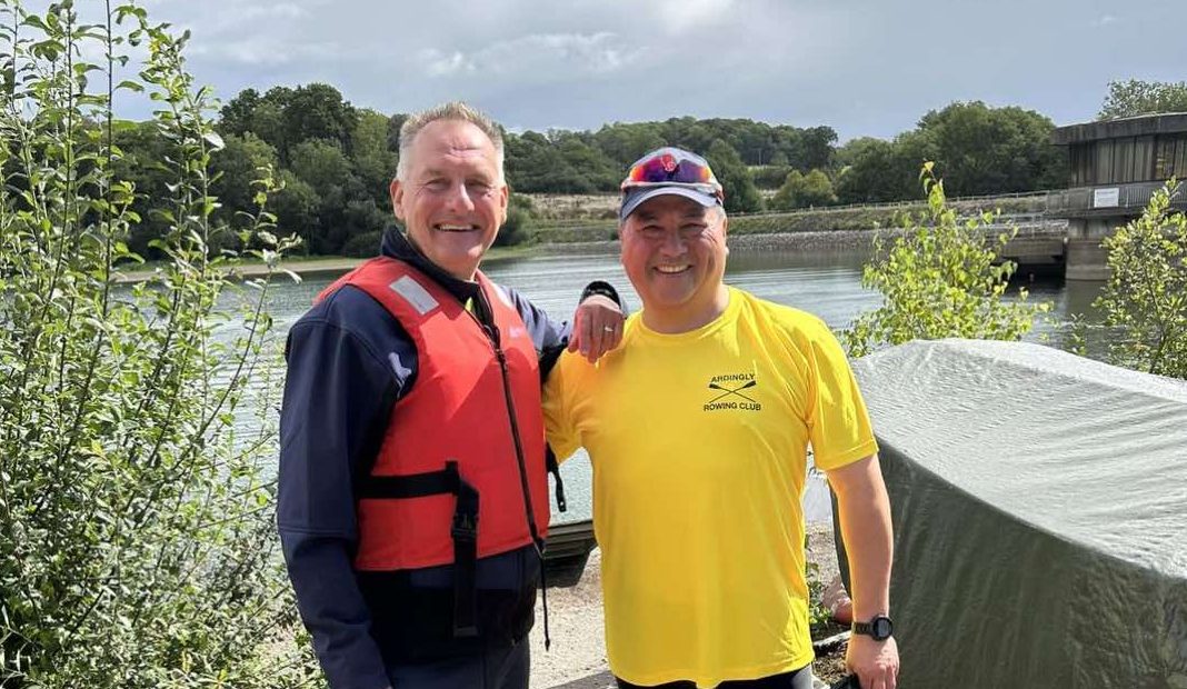 Two smiling men beside a lake. Rowing coach wears red life jacket. Athlete masters rower in yellow tshirt.