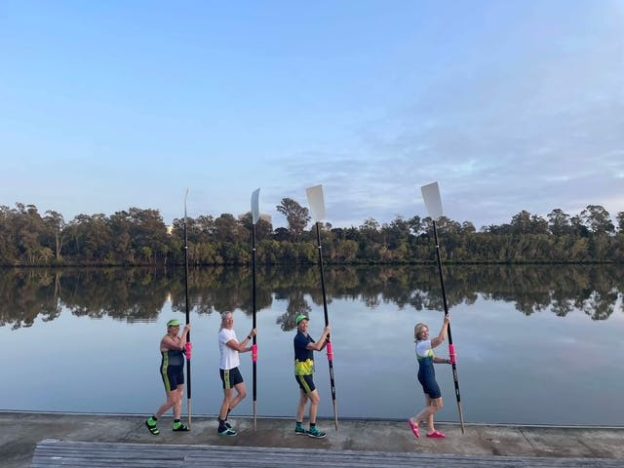 Four women holding rowing oars upright beside a river