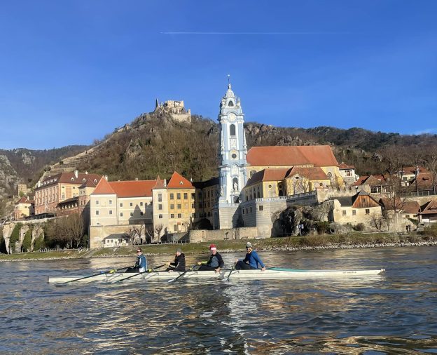 Mens quadruple scull on a river in front of a German church and old houses